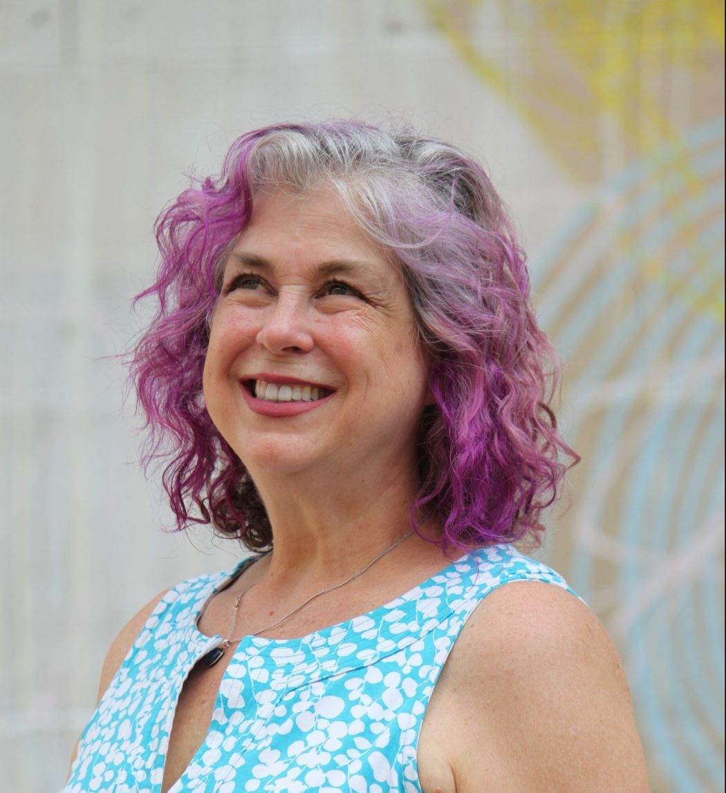 Debra looking up and smiling with a blue shirt and hair dyed purple