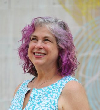 Debra looking up and smiling with a blue shirt and hair dyed purple