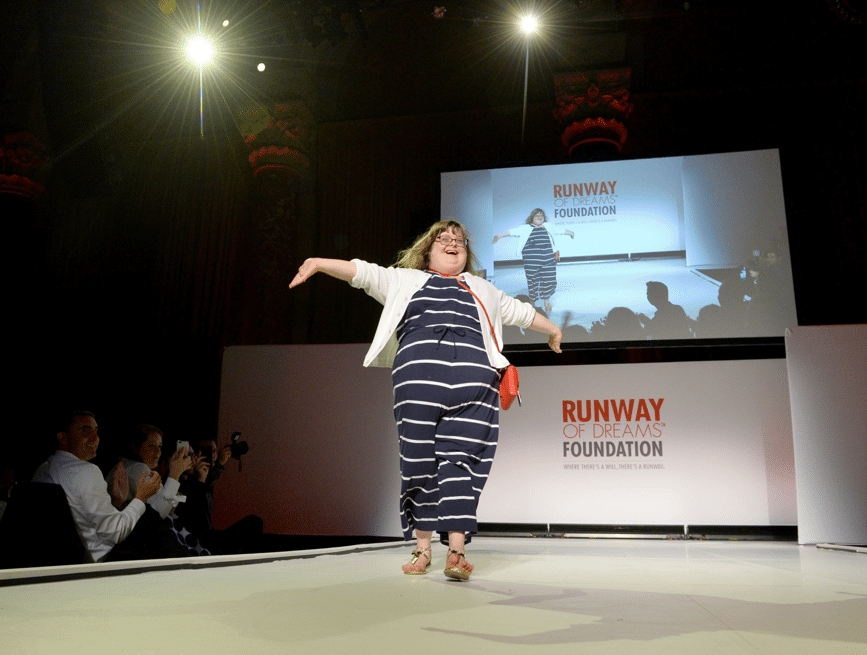 Sara walking down the fashion runway, arms in the air, wearing a navy blue striped jumpsuit and white sweater