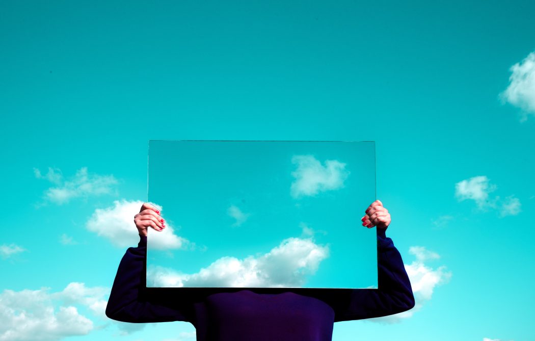 woman holding a mirror in front of her face, reflecting the sky and clouds