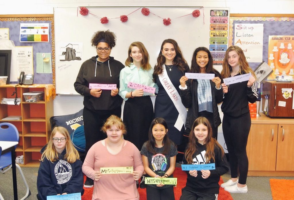 Amanda with a group of eight middle school girls in a classroom. Amanda wears her Miss Great Lakes sash. The girls hold up colored paper with their superpowers written on them.