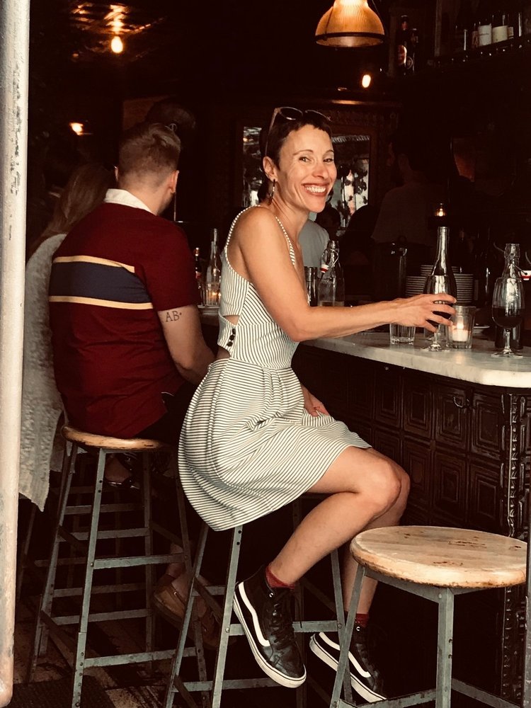 Emily smiles, sitting on a bar stool wearing a sun dress and sneakers