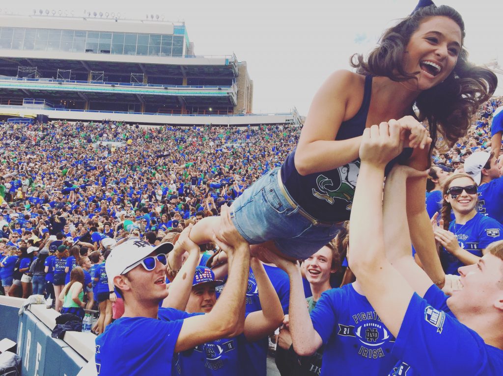 Amanda body surfing at a football game. She is wearing a jean skirt, a blue Notre Dame tank top and is laughing. A large stadium filled with football fans is behind her.