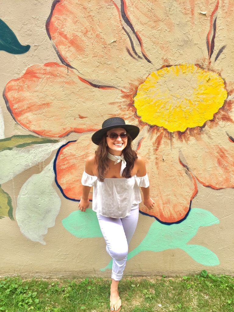 Amanda smiles and poses in front of a wall painted with a mural of a large flower. She wears white pants, a white off the shoulder blouse and a stylish sun hat. 
