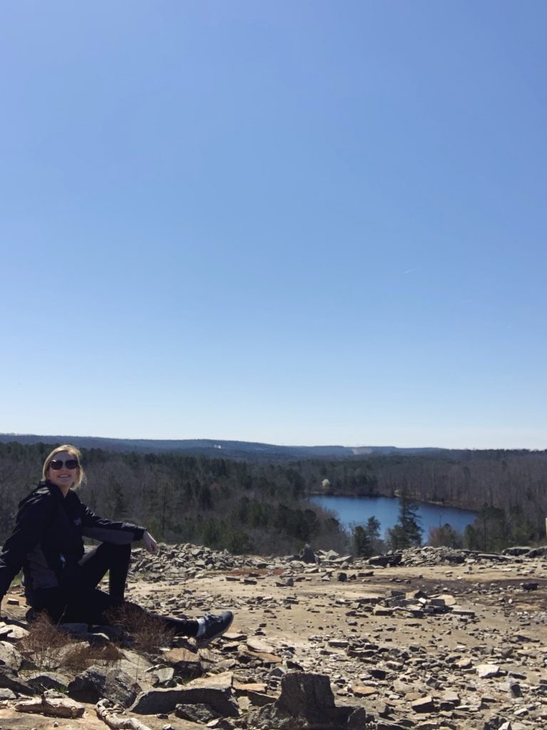 Mackenzie in athletic clothing, sitting at the top of a mountain, with a lake and forest in the background.