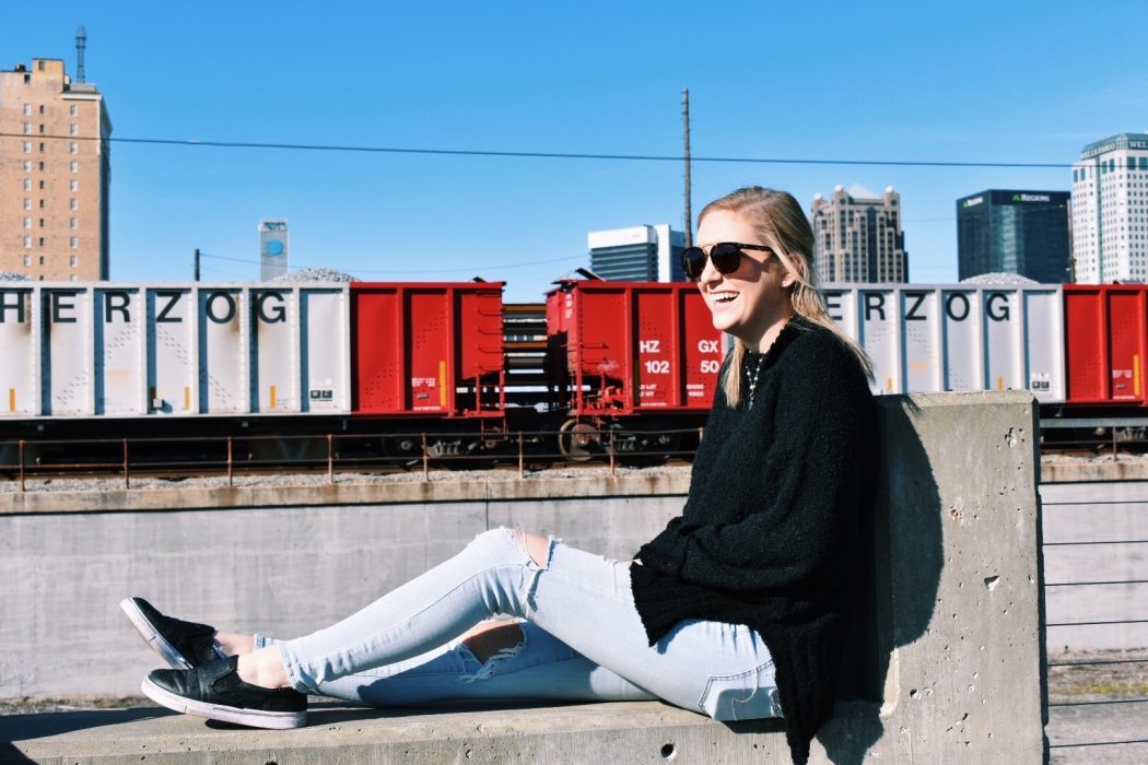 MacKenzie smiles sitting on a concrete bench wearing sunglasses