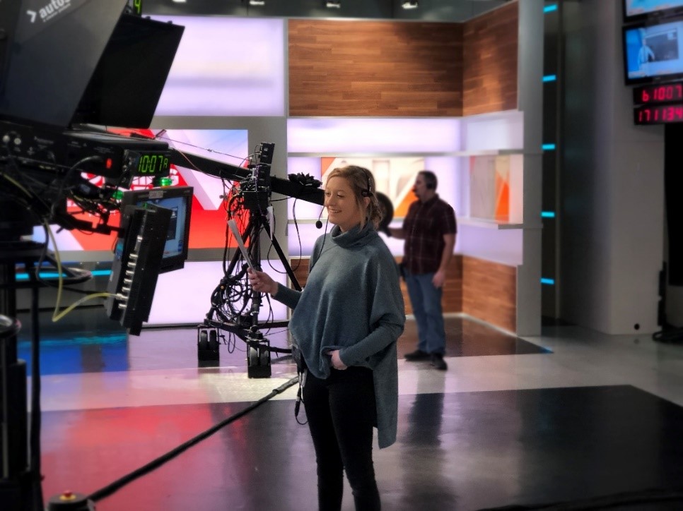 Mackenzie on the floor of a newsroom behind camera equipment looking at playback on a monitor. She is wearing a headset and smiling.