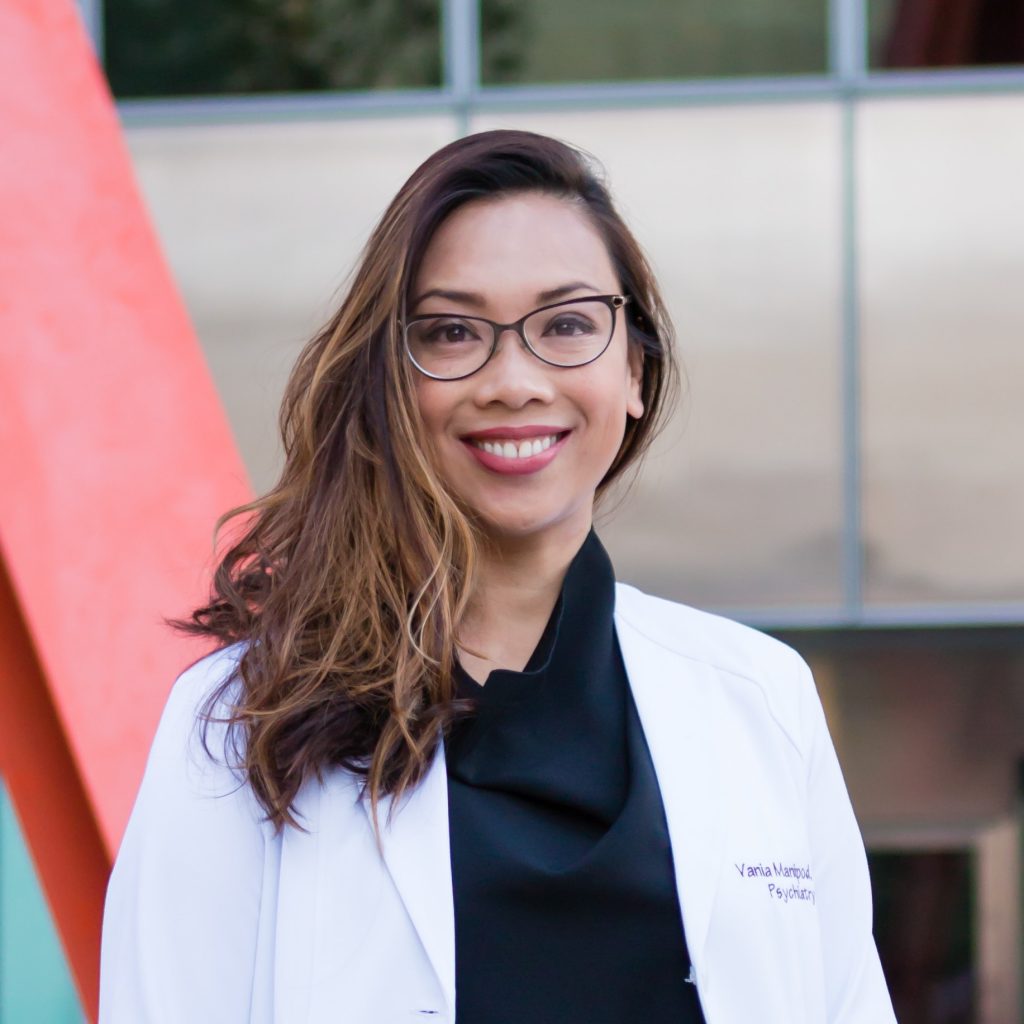 Dr Vania smiles wearing a white medical coat and glasses