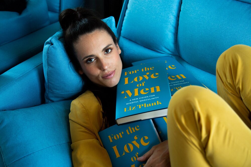 Liz Plank in yellow suit on blue couch, holding copies of her book titled for the love of men.