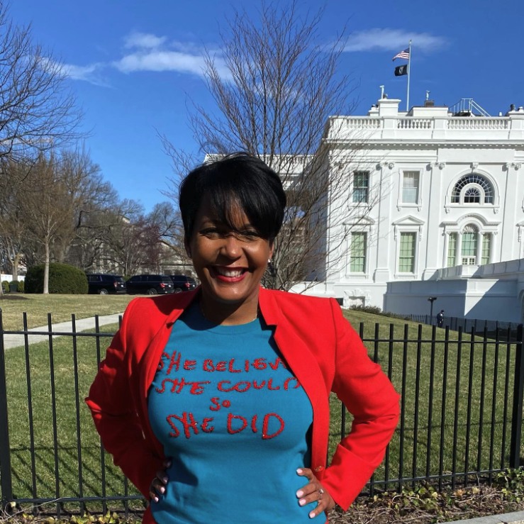Keisha smiling in front of the White House, wearing a red suit jacket and blue shirt that reads "She believed she could, so she did"
