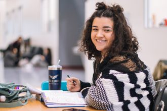 Teenage girl with curly hair, sitting at desk in school, smiling. Textbook open in front of her