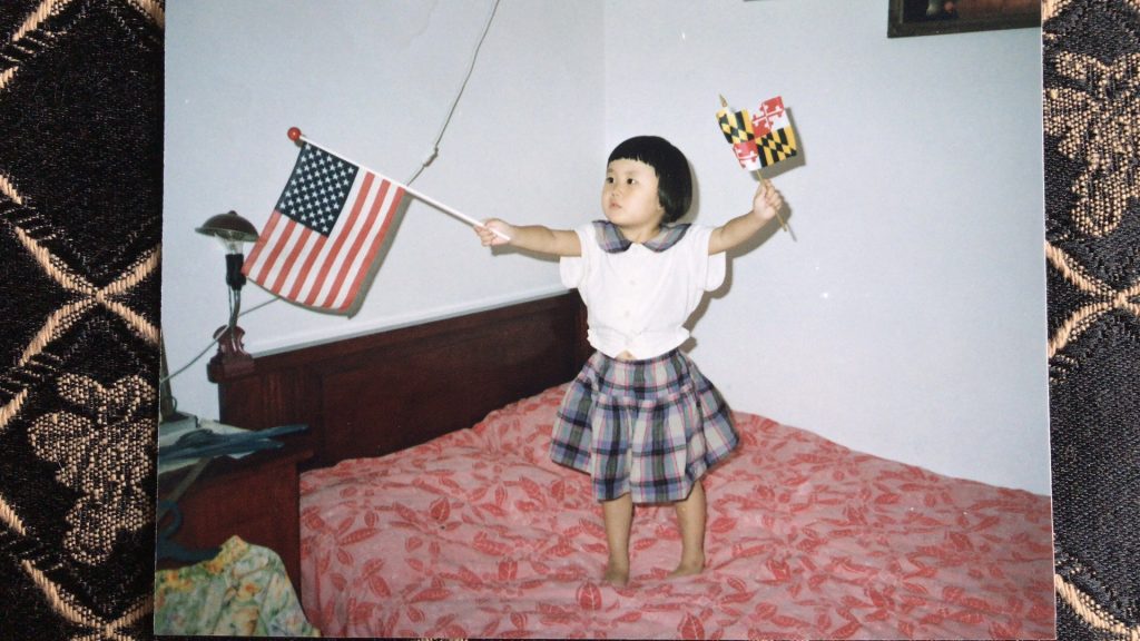 4 year old Lind standing on a bed holding an American flag in one hand