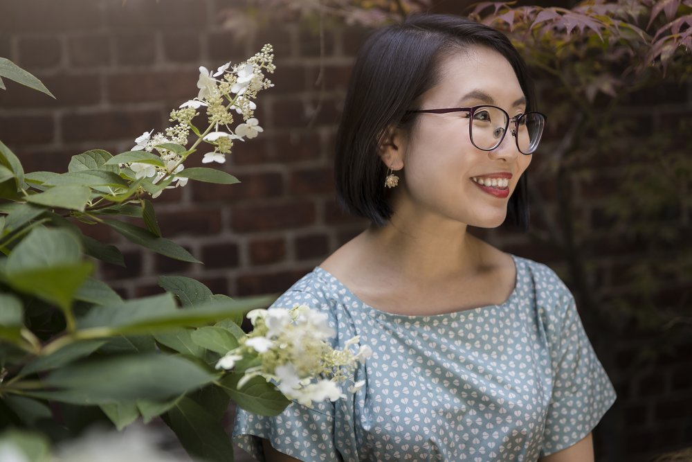 Linda Yi smiling and looking off camera, in front of a brick wall and flowers