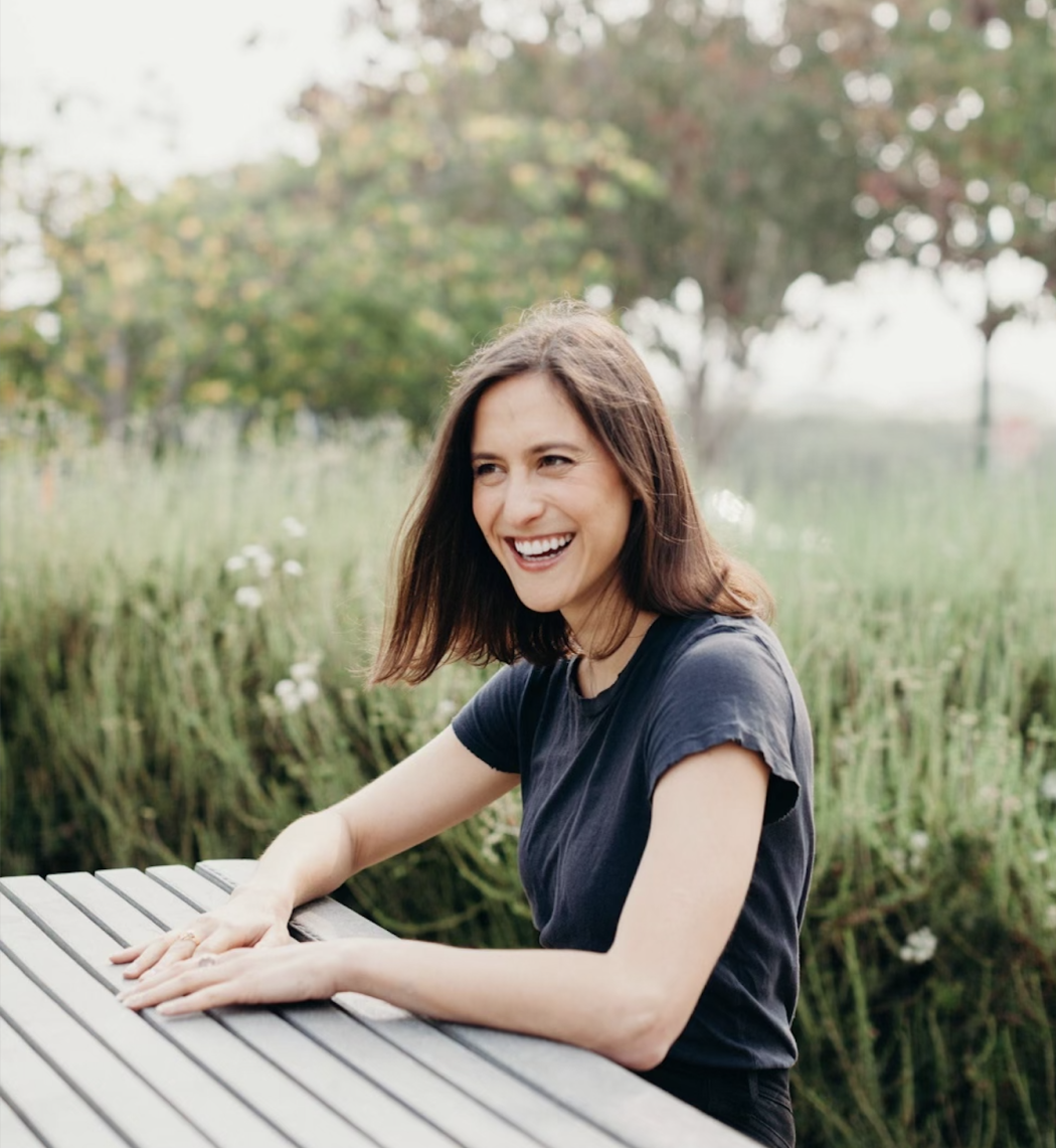 Emily smiling sitting at a picnic table in a lush meadow.